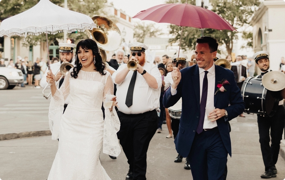 Newlywed couple in wedding parade with band, parasols, fringe wedding dress, and navy blue wedding suit.